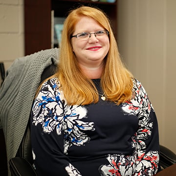 green_cassie Woman with long read hair smiling into camera wearing black floral shirt.