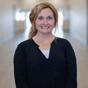 Woman with blonde hair pulled into ponytail smiling into camera while wearing black long sleeved shirt.