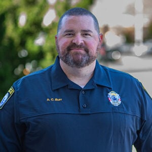 Man with buzzed dark hair and beard smiling into camera wearing shirt with badge on.