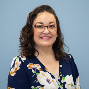 A woman with long curly black hair and glasses is wearing a black floral dress.