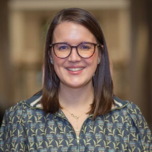A woman with shoulder length brunette hair and glasses is wearing a green and yellow floral shirt while smiling.