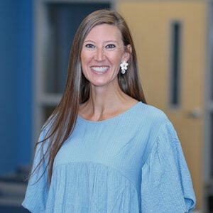 A woman with long brunette hair is wearing silver earrings and a baby blue top while smiling at the camera.