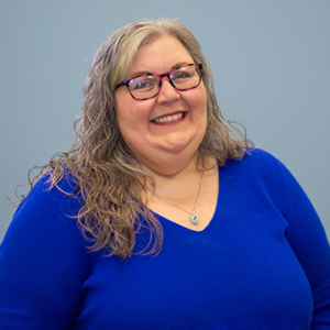 Woman with salt and pepper hair smiling into camera wearing royal blue shirt.