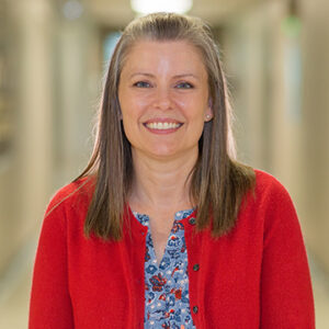 A woman with long brunette hair is wearing a blue floral shirt and a red cardigan sweater while smiling at the camera.