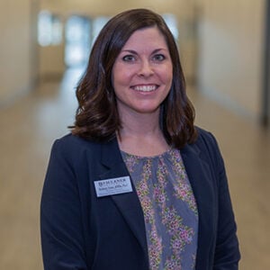 A woman with short wavy brunette hair is wearing a light blue floral shirt and a black blazer while smiling.