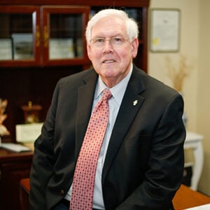 Man with short white hair wearing glasses and smiling into camera while wearing black suit with red tie.