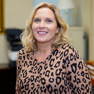 Woman with mid length blonde hair smiling into camera wearing leopard print shirt.