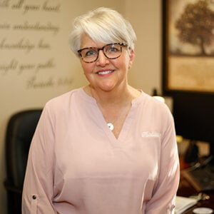 Woman with short white hair wearing glasses and a pink shirt smiling into the camera.