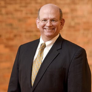 Man wearing glasses and dark suit with yellow tie smiling into camera.