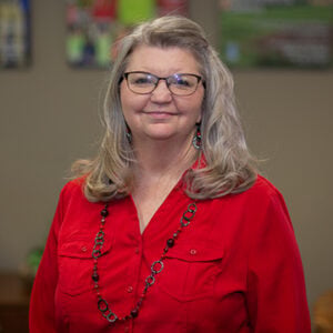 A woman with shoulder length silver hair is wearing a red button up shirt and a ringlet necklace with glasses while smiling into the camera.