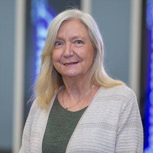 A woman with white hair is wearing an olive green shirt and a gray cardigan sweater while smiling at the camera.