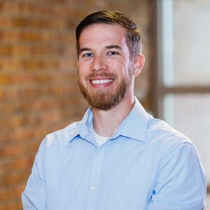 A man with brown hair and a brown beard is wearing a light blue dress shirt while smiling at the camera.