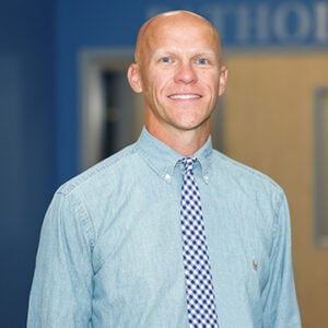 A man wearing a light blue dress shirt and a white and navy blue checkered tie is smiling.