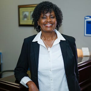 A woman with short curly hair is smiling at the camera and wearing a white dress shirt and a black blazer with the sleeves pulled up.