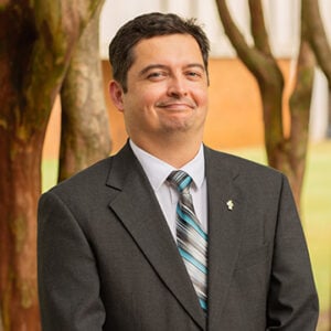Man with short dark hair smiling into camera wearing dark suit and multi coloured tie.