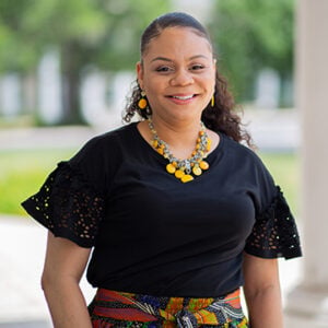 A woman with dark curly hair slicked back wearing a black shirt, a yellow beaded necklace, and matching yellow earrings is smiling into the camera.