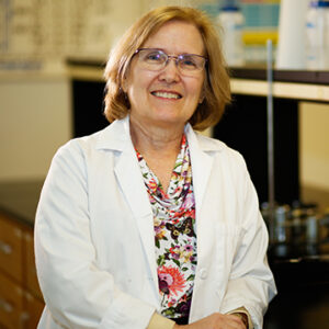 A woman with shoulder length blonde hair and glasses is smiling at the camera while wearing a floral shirt and a white lab coat.