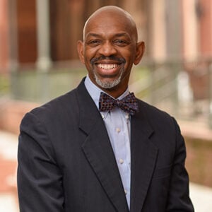 A man with a salt and pepper goatee wearing a light blue dress shirt, a black blazer, and a bowtie.