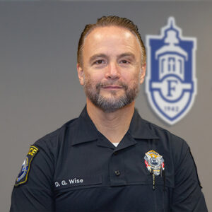 A man with short brunette hair wearing a black shirt with his name and badge is smiling.