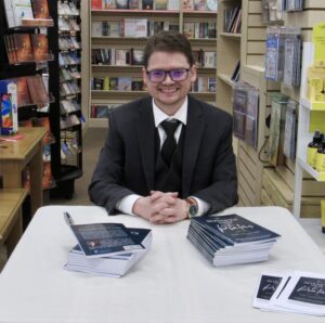 A man with short brown hair and glasses is wearing a black suit with a black tie smiling while sitting at a table covered in books.