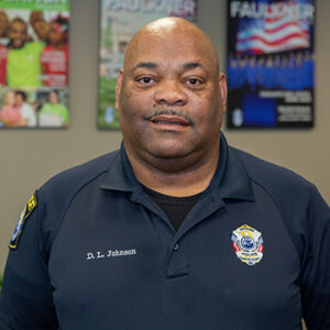 A man with a salt and pepper mustache is wearing a black shirt with his name and badge on it.