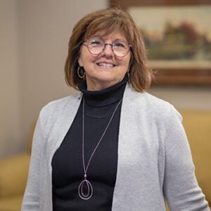 A woman with mid length brunette hair is smiling into the camera wearing glasses, a black shirt, a grey cardigan sweater, and a silver necklace.