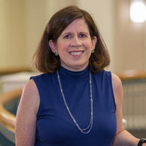 A woman with shoulder length brunette hair is wearing a silver necklace and a navy blue dress while smiling at the camera.