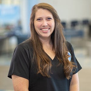 A woman with long brunette hair is wearing black Faulkner Health Sciences scrubs and is smiling at the camera.