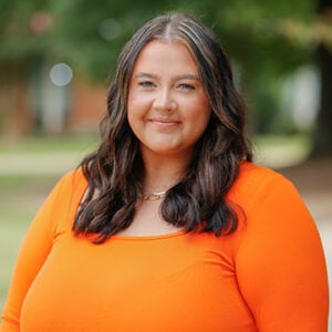 Woman with long dark hair smiling into camera wearing orange outfit.