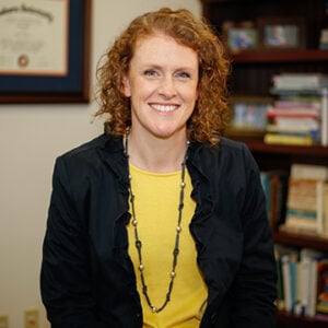 A woman with curly red hair is wearing a black and pearled necklace, a yellow shirt, and a black blazer while smiling at the camera.