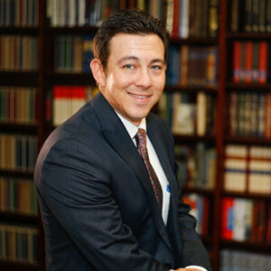 A man with dark brunette hair is wearing a white dress shirt, a black blazer, and a tie while smiling at the camera.