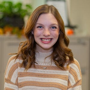 A woman with shoulder length brunette hair is wearing a white and orange striped sweater while smiling.