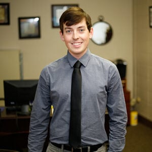 A man with short brunette hair is wearing a gray button up with a black tie while smiling.