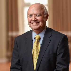 A man in a light blue dress shirt, a black blazer and a yellow tie is smiling into the camera.