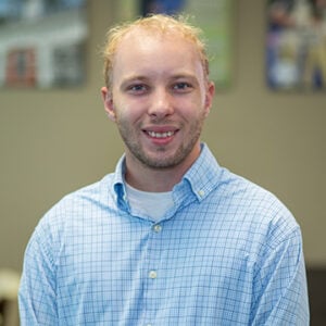 A man with short blonde hair is wearing a white and blue checkered dress shirt while smiling.