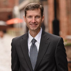 A man with short brown hair is wearing a white dress shirt, a black blazer, and a blue tie while smiling.