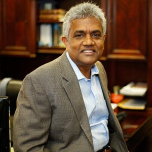 A man with salt and pepper hair is wearing a light blue dress shirt and a light brown blazer while smiling.