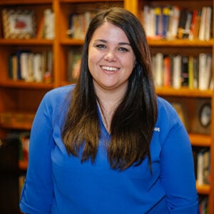 A woman with long brunette hair is wearing a royal blue shirt while smiling.