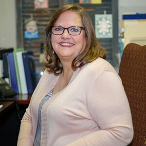 A woman with shoulder length brunette hair is wearing glasses, a black and white striped shirt, and a pink cardigan while smiling.
