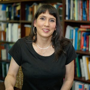 A woman with long black hair is wearing a silver necklace and a black top while smiling.