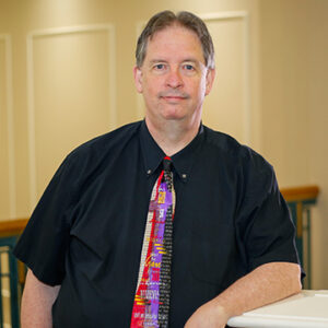 A man with short black hair is wearing a black dress shirt with a multi-colored tie and smiling.