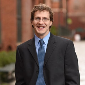 A man with short curly brown hair is wearing glasses, a light blue dress shirt, a navy blue tie, and a black blazer while smiling.