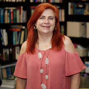 A woman with mid length red hair is wearing a necklace and a pink shirt while smiling.