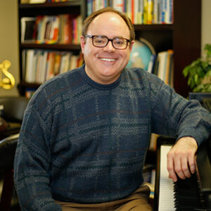 A man with short brunette hair is wearing glasses and a blue sweater while smiling.