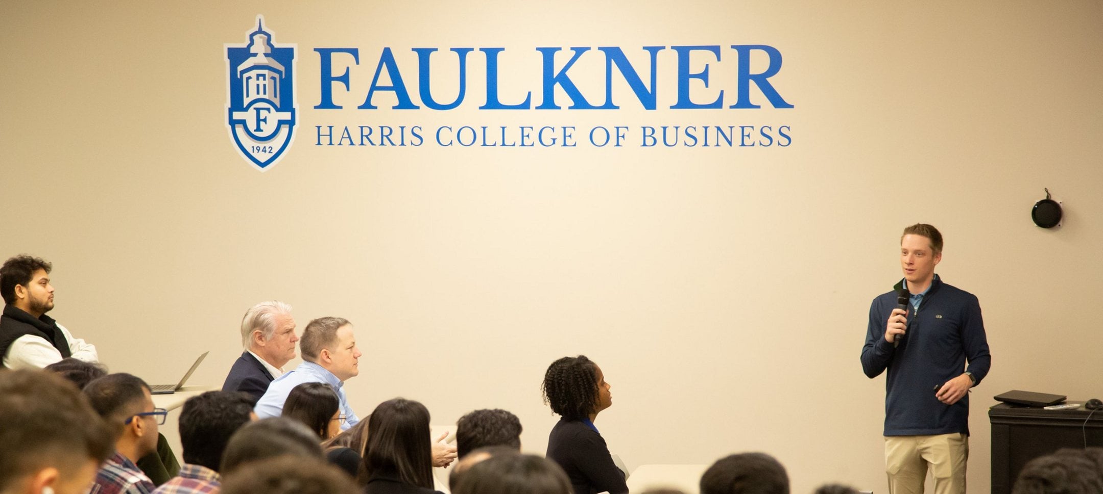 Speaker presenting to students and faculty beneath the Faulkner Harris College of Business logo