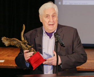 Joe Beam, speaks into a microphone while holding his Alumnus of the Year award, a bronzed eagle in flight.