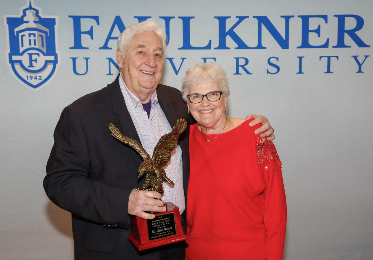Joe Beam poses with his wife, Alice Beam in front of a Faulkner backdrop while holding his Alumnus of the Year Award.