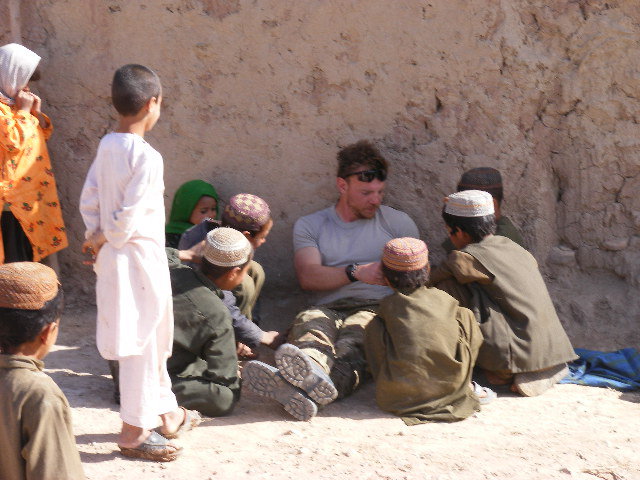 SSG Michael Hosey sits against a stone wall surrounded by several Afghan children as he talks with them during one of his combat tours. 