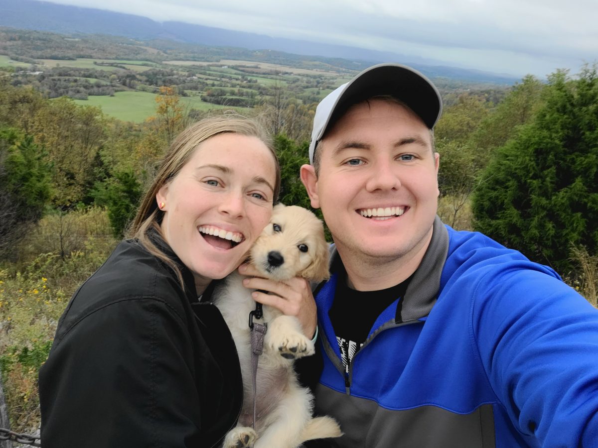 Elliott Graves, right poses with his wife, Tia and pet golden doodle, Ahsoka in front of a mountain landscape.