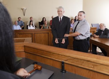 Handcuffed defendant stands with bailiff in courtroom